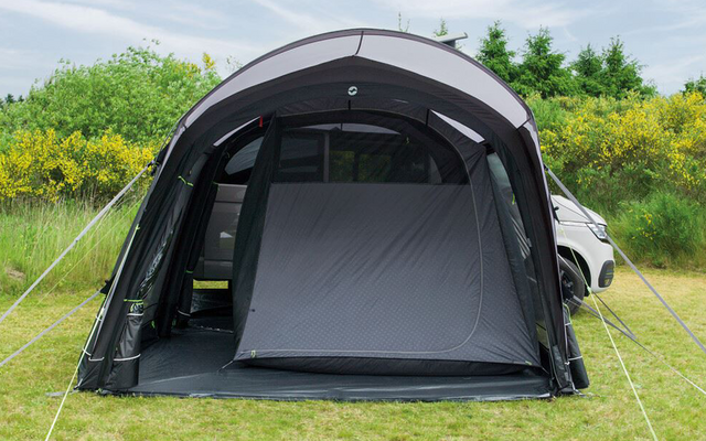 Dunkelgraues aufblasbares Vorzelt mit geschlossener Innenzeltkammer, vordere Öffnung sichtbar; rechts teilweise ein weißes Auto; Grasfläche, gelb blühende Büsche im Hintergrund und bewölkter Himmel