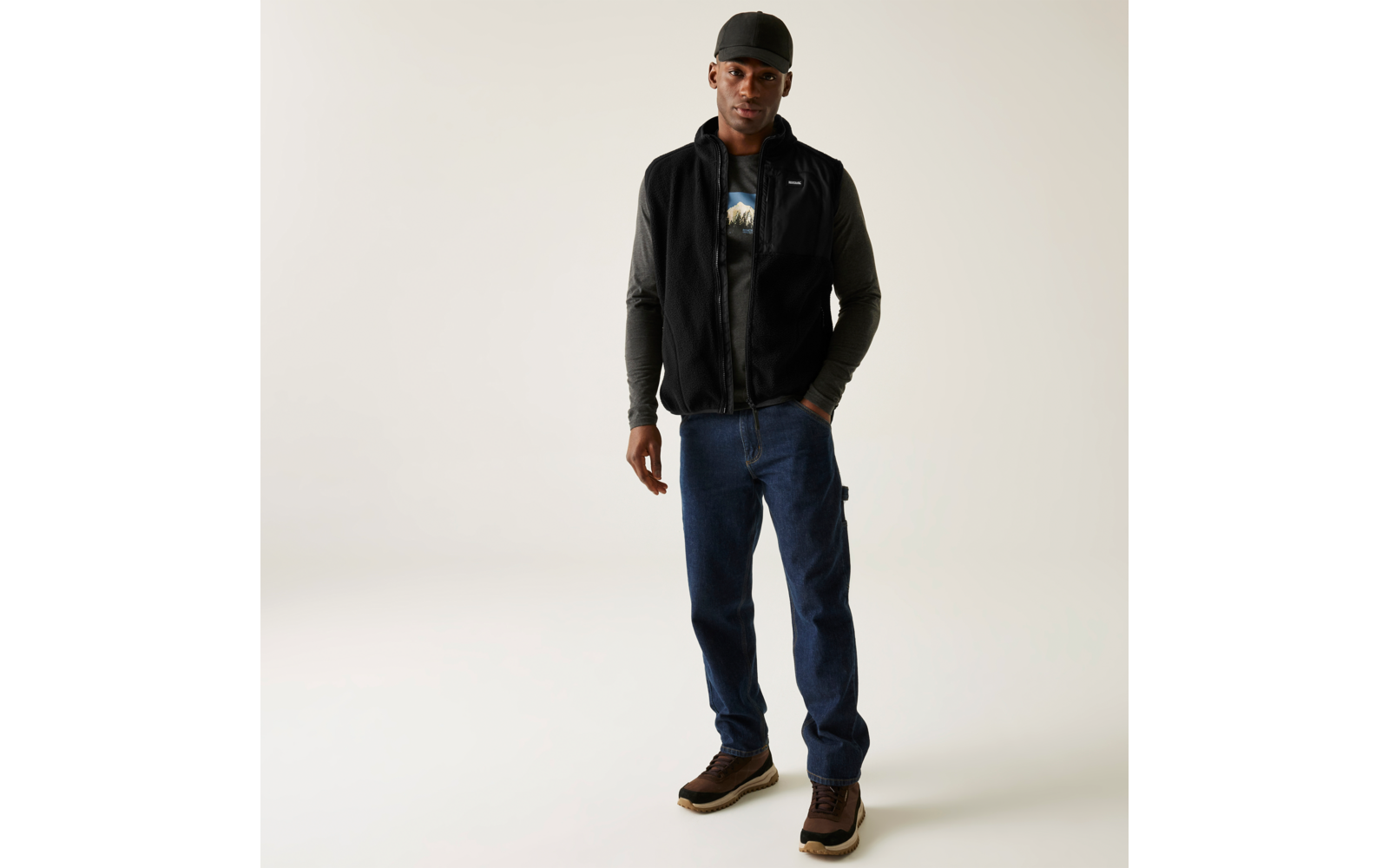 Mann in schwarzer Jacke, blauer Jeans und braunen Schuhen steht vor heller Wand