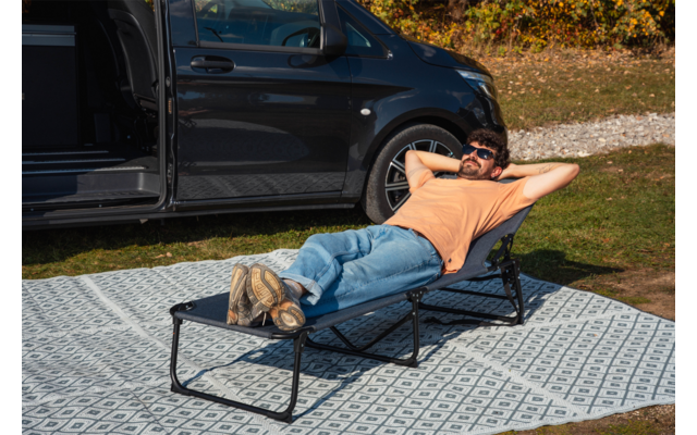 Person liegt auf einer zusammenklappbaren Liege auf einem gemusterten Picknickteppich neben dem offenen Türbereich eines dunklen Vans, trägt orangefarbenes T-Shirt, Jeans und Sonnenbrille, Hände hinter dem Kopf, Gras und Herbstlaub im Hintergrund