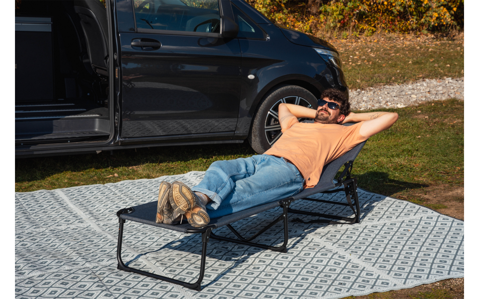 Person liegt auf einer zusammenklappbaren Liege auf einem gemusterten Picknickteppich neben dem offenen Türbereich eines dunklen Vans, trägt orangefarbenes T-Shirt, Jeans und Sonnenbrille, Hände hinter dem Kopf, Gras und Herbstlaub im Hintergrund