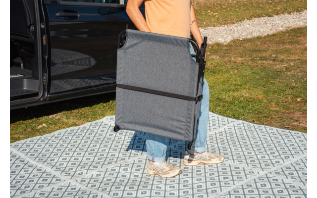 Person von der Taille abwärts trägt zusammengeklappten grauen Klappstuhl neben geöffneter Autotür, steht auf gemustertem Outdoorteppich auf Gras