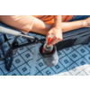 Arm einer sitzenden Person mit orangefarbenem T‑Shirt und Jeansshorts; Hand hält eine Glasflasche mit roter Flüssigkeit in einem grauen Getränkehalter, der an der Armlehne eines Klappstuhls befestigt ist; darunter ein blauer Teppich mit geometrischem