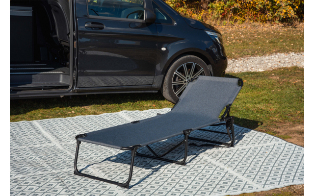 Dunkler Van mit geöffneter Schiebetür, davor eine graue Liege auf einem gemusterten Outdoor-Teppich, Rasen und Laub im Hintergrund