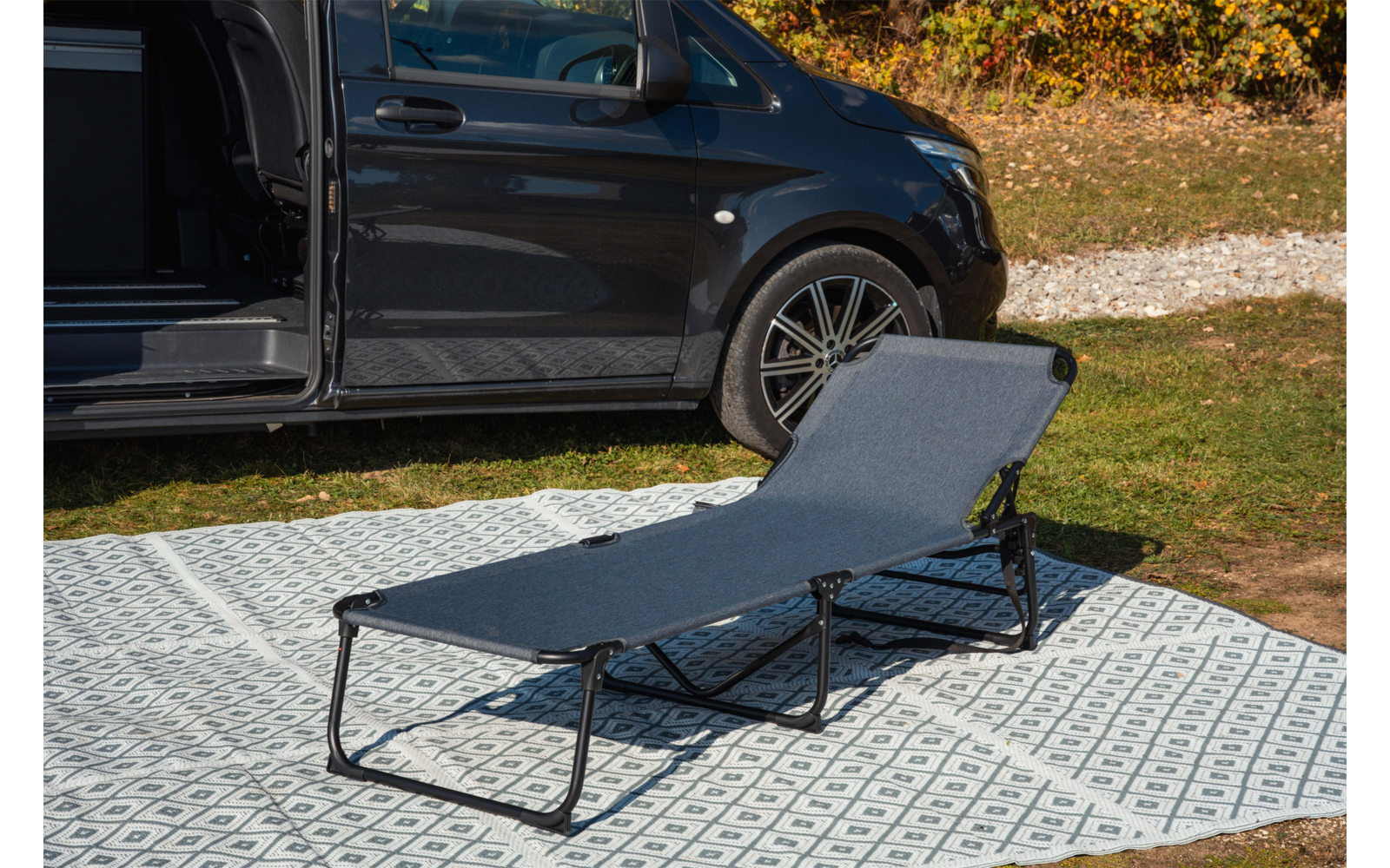 Dunkler Van mit geöffneter Schiebetür, davor eine graue Liege auf einem gemusterten Outdoor-Teppich, Rasen und Laub im Hintergrund