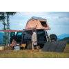 Femme debout à côté d'une voiture avec une tente sur le toit ouverte, équipement de camping sur une pelouse