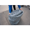 Person in blauem Sweatshirt und schwarzen Hosen hält einen offenen grauen Sack mit aufgerolltem grauen Material; Asphalt und Gehweg sichtbar