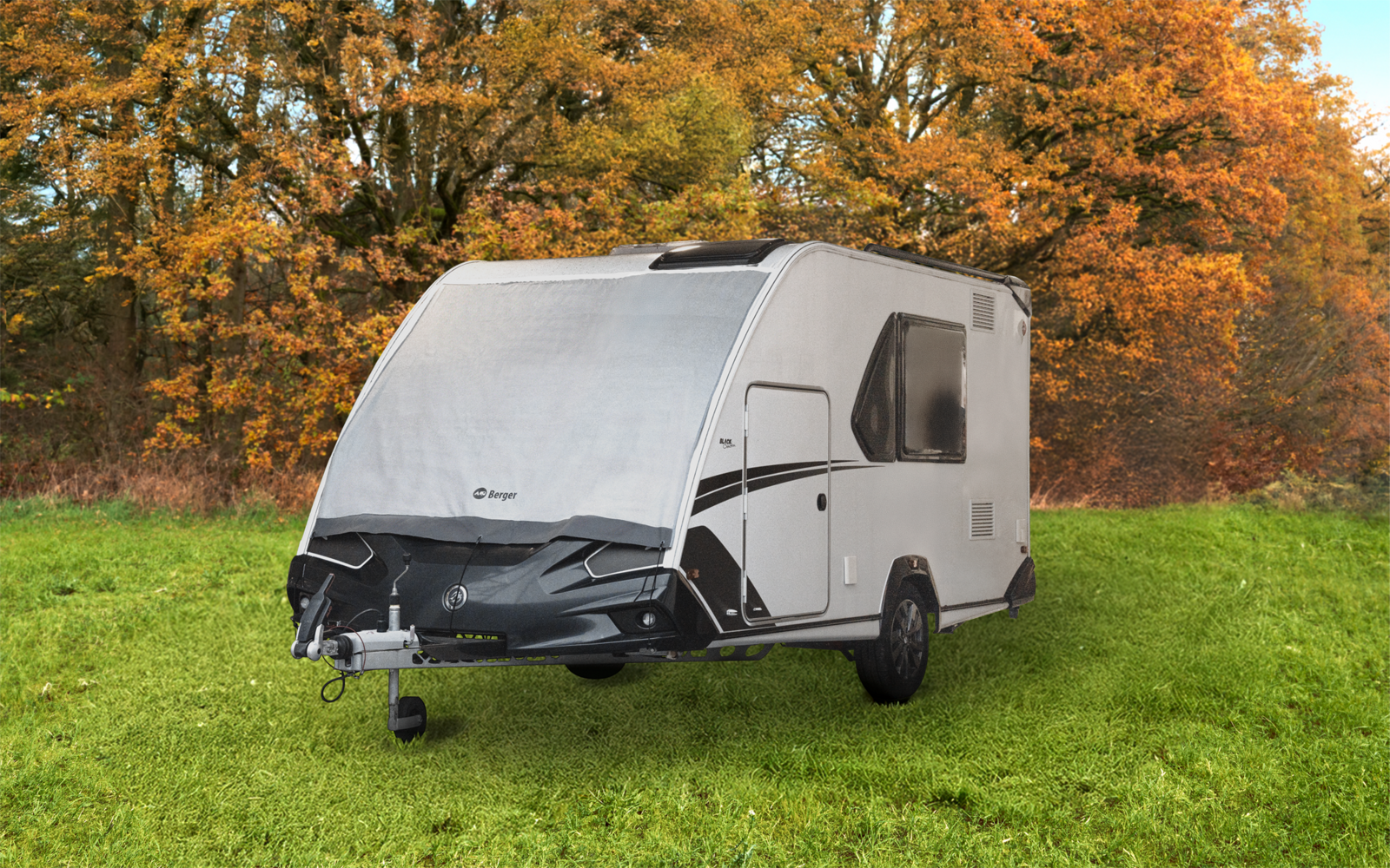 Weißer Wohnwagen auf grüner Wiese, Anhängerkupplung und Rad im Vordergrund, herbstlich gefärbte Bäume im Hintergrund