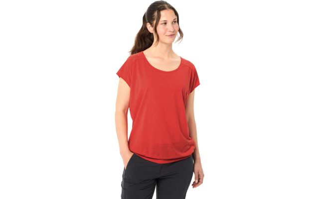 Personne aux cheveux bruns attachés en queue de cheval, portant un haut rouge à manches courtes et un pantalon foncé, mains dans les poches, regard vers la droite, fond blanc