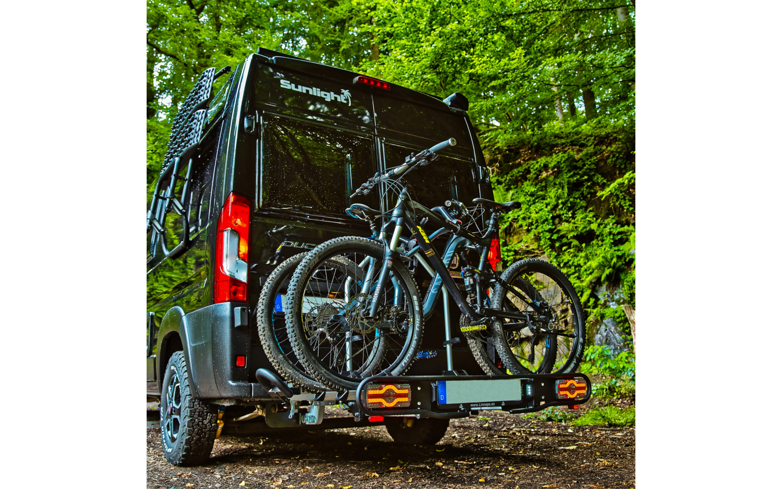Black camper van with 'Sunlight' logo; rear bike rack holding two mountain bikes; green forest in the background; dirt ground