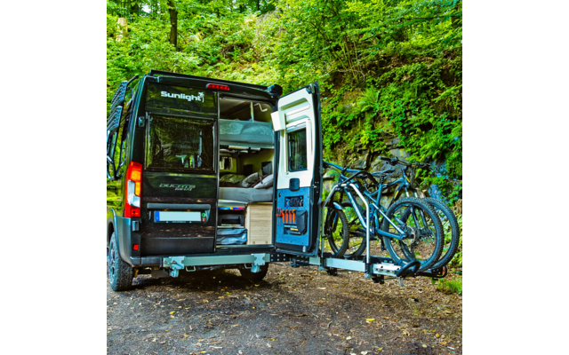 Black camper van with rear doors open showing bed and storage, three bicycles on a rear rack, forested background