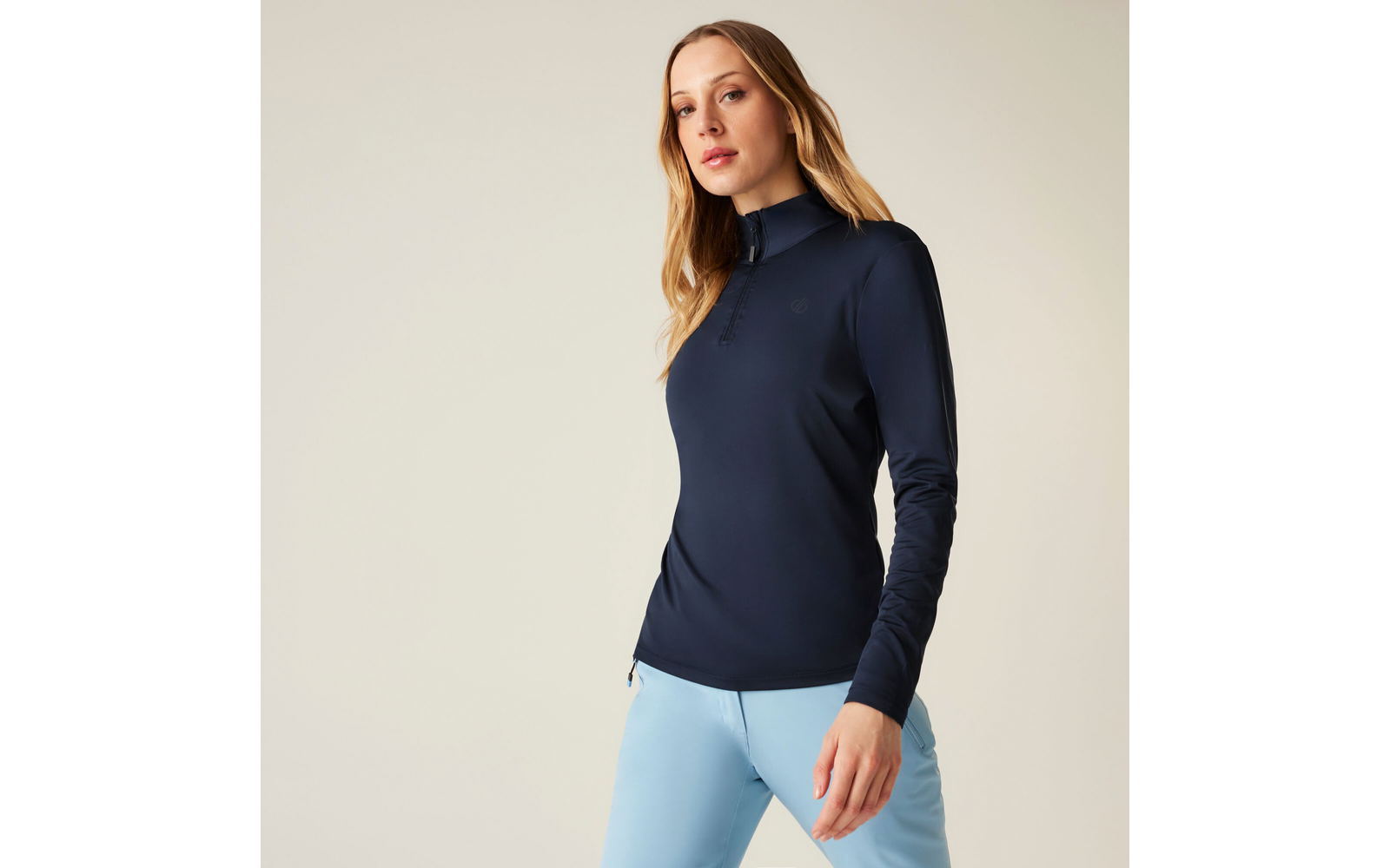 Femme aux longs cheveux blonds portant un pull bleu foncé et un pantalon bleu clair devant un fond clair