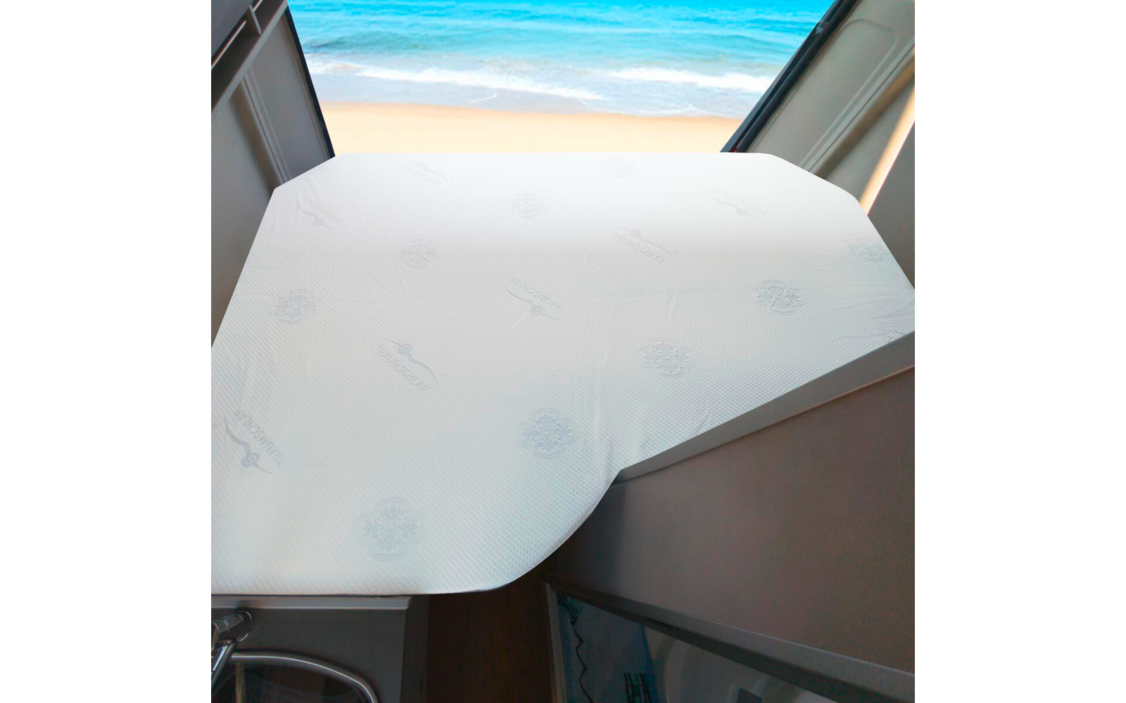 Weißer, strukturierter Tischbezug auf einer eckigen Tischfläche vor einem offenen Fenster mit Sandstrand und Meer; metallisches Tischbein und Teil eines Schranks sichtbar