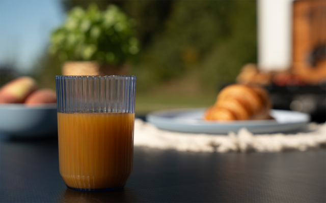 Geripptes Glas mit Orangensaft auf dunklem Tisch, im Hintergrund unscharf Teller mit Croissants, Schale mit Obst und Pflanzen