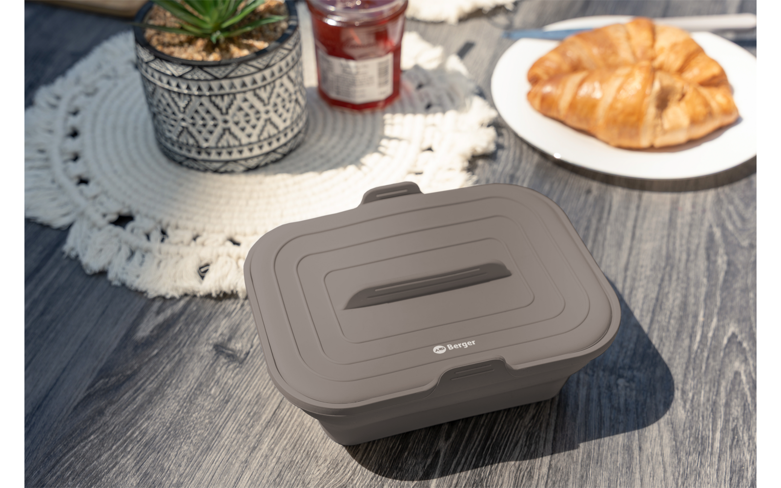 Geschlossene graue Kunststoffbox mit Deckel und Griff auf einem Holztisch; im Hintergrund ein Topf mit Sukkulente auf einem Häkeldeckchen, ein Marmeladenglas und ein Teller mit Croissant