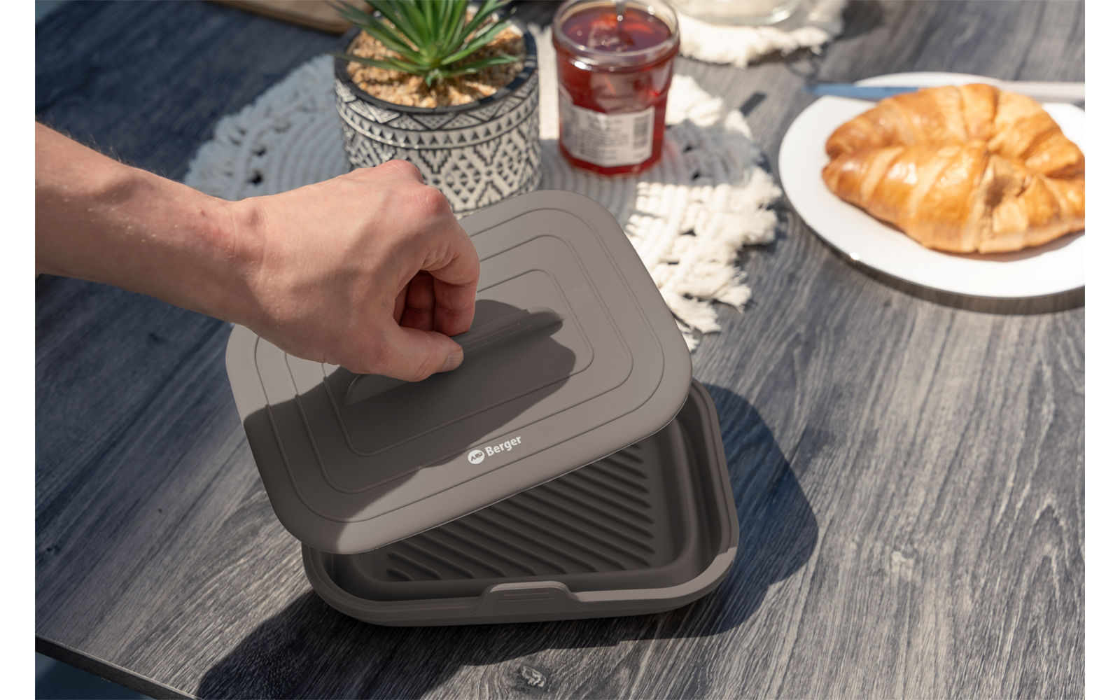 Hand hebt den Deckel eines grauen, quadratischen, zusammenklappbaren Silikonbehälters auf einem Holztisch; daneben ein kleiner Topf mit Sukkulente, ein Marmeladenglas und ein Teller mit Croissant