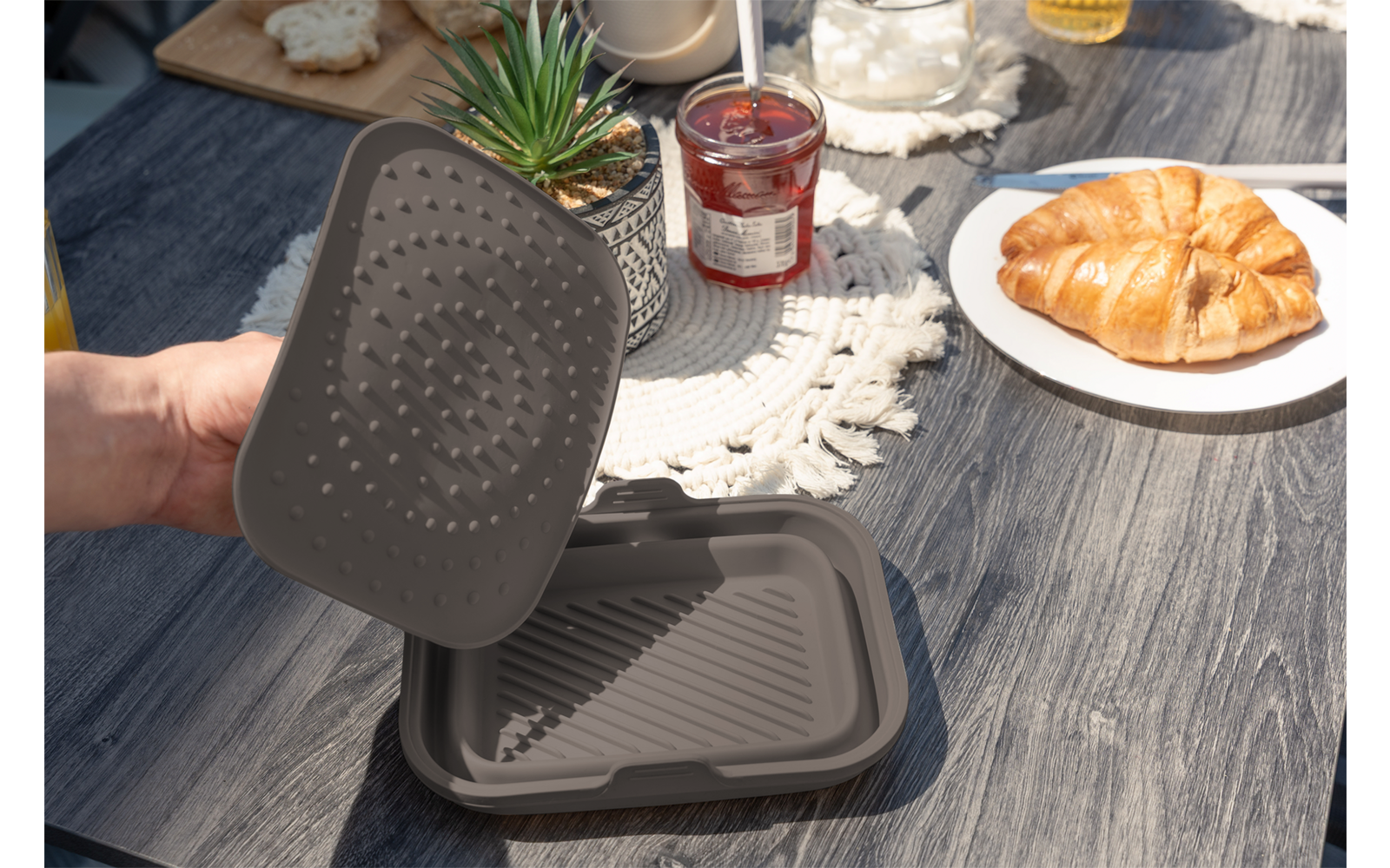 Eine Hand hält einen grauen, rechteckigen Deckel über einem passenden Behälterboden auf einem Holztisch; daneben ein Croissant auf einem weißen Teller, ein Glas mit roter Marmelade, ein Wasserglas, eine kleine Topfpflanze und ein gehäkelter Untersetz