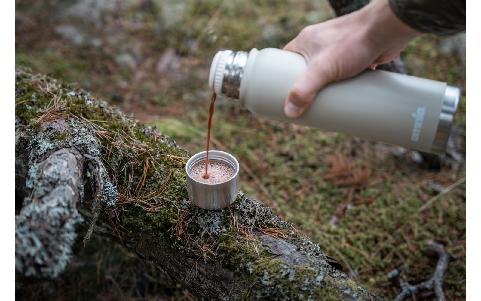 Hand giet donkere vloeistof uit een thermosfles in een klein metalen bekertje op een met mos bedekte boomstam
