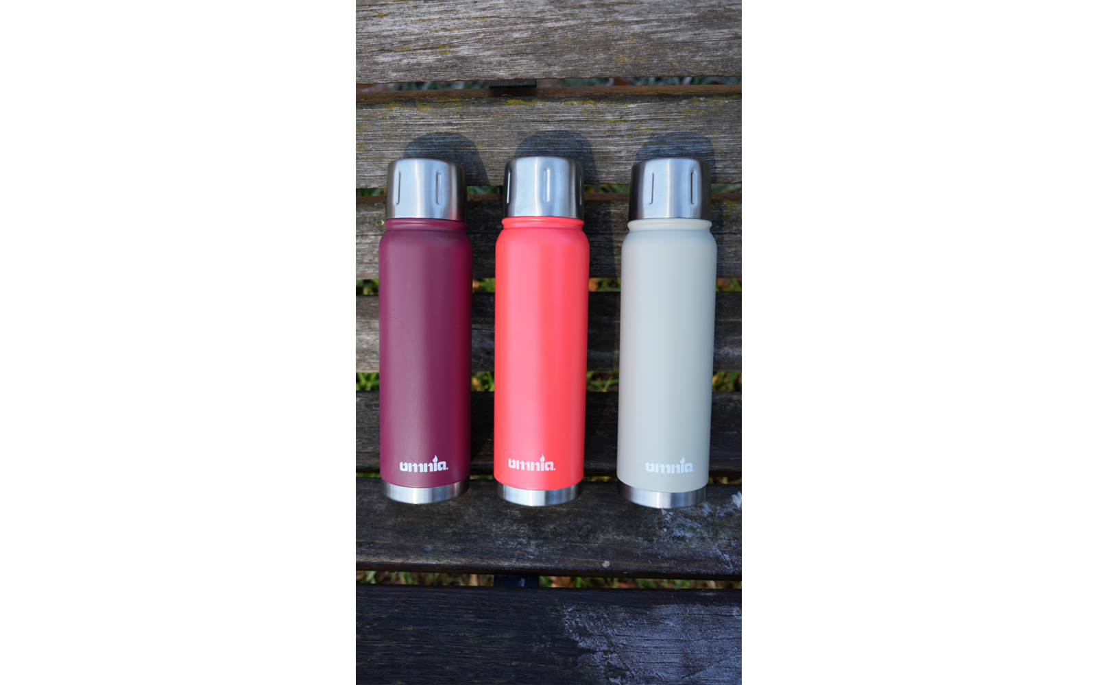 Drie hoge flessen met schroefdoppen in bordeaux, koraal en lichtgrijs naast elkaar op een houten bank