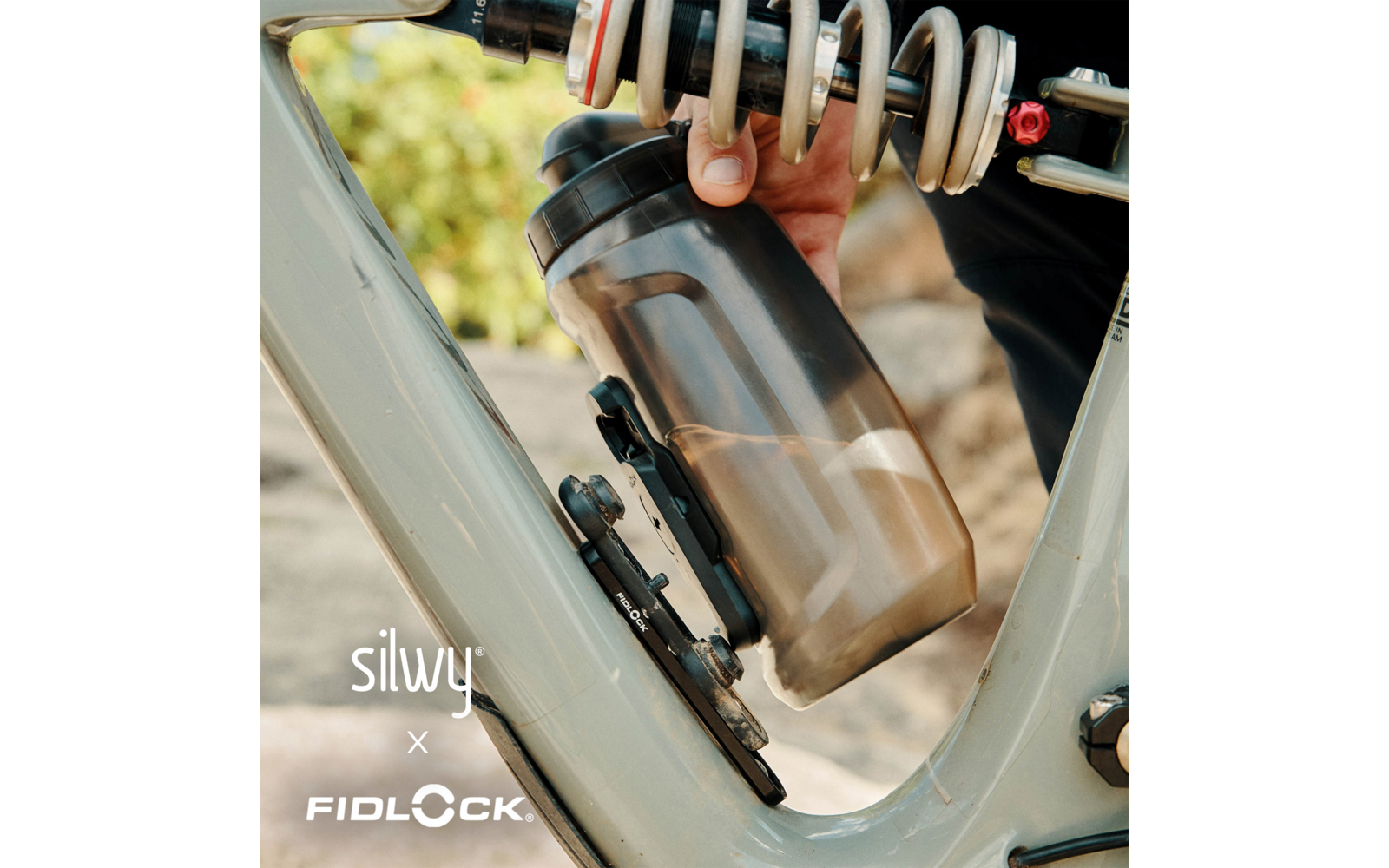 Hand, die eine transluzente Trinkflasche in einen am grauen Fahrradrahmen montierten Flaschenhalter steckt; Fahrradfeder und Logos 'silwy x FIDLOCK' sichtbar