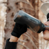 Nahaufnahme einer Person, die aus einer grauen Trinkflasche mit Aufschrift silwy + FIDLOCK trinkt; Teil des Gesichts, Sonnenbrille, helle Kappe und schwarze Handschuhe sichtbar; unscharfer Waldhintergrund
