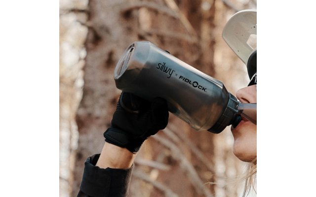 Nahaufnahme einer Person, die aus einer grauen Trinkflasche mit Aufschrift silwy + FIDLOCK trinkt; Teil des Gesichts, Sonnenbrille, helle Kappe und schwarze Handschuhe sichtbar; unscharfer Waldhintergrund
