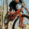 Person in der Luft mit orangefarbenem Mountainbike zwischen Bäumen; sichtbare Knieprotektoren, Hände am Lenker, Vordergabel mit FOX-Logo