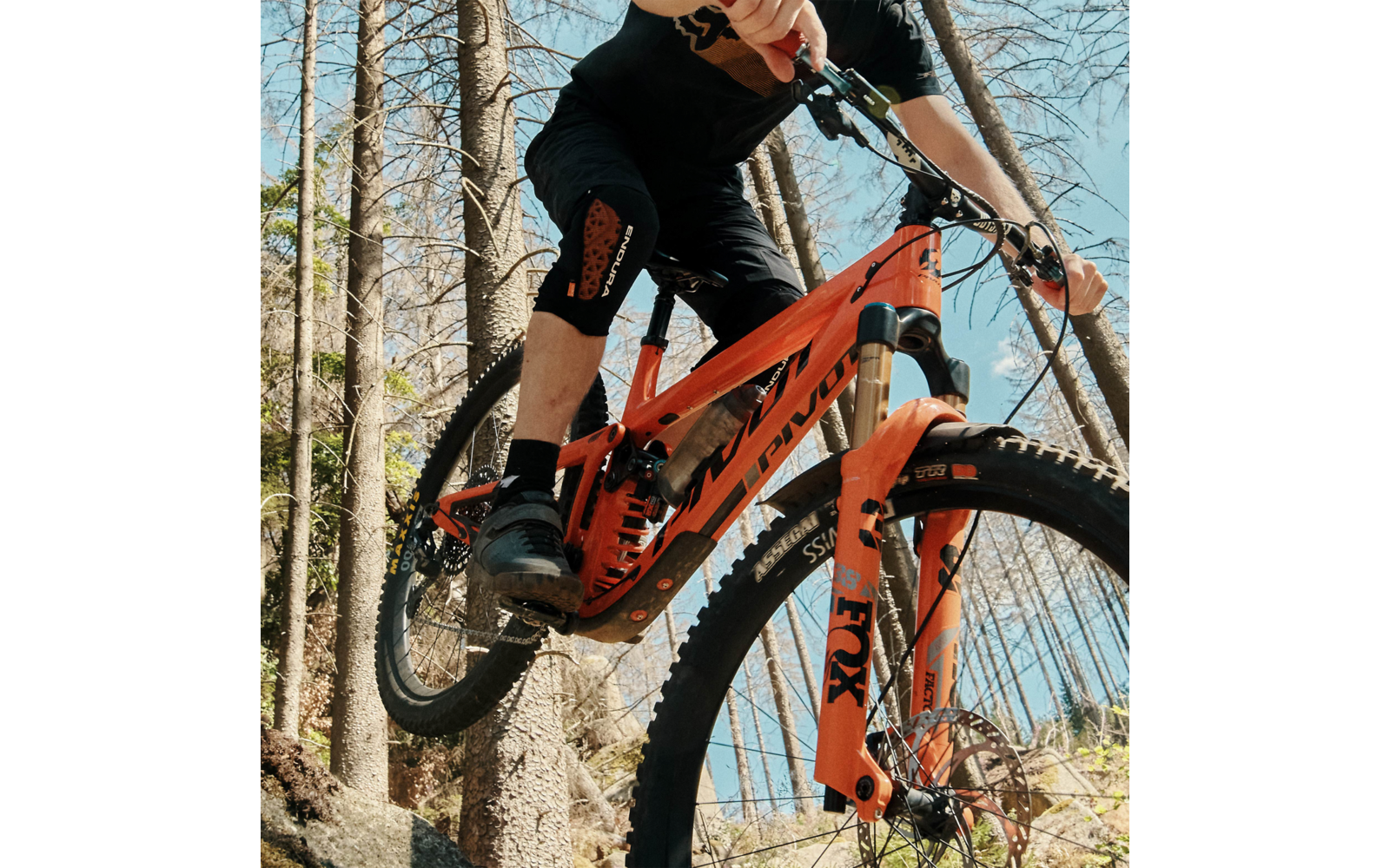 Person in der Luft mit orangefarbenem Mountainbike zwischen Bäumen; sichtbare Knieprotektoren, Hände am Lenker, Vordergabel mit FOX-Logo