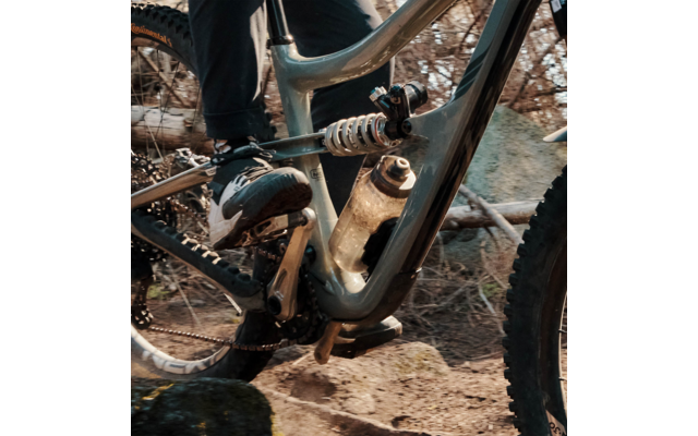 Nahaufnahme eines Mountainbike-Rahmens mit Spiralfederdämpfer, Trinkflasche im Rahmen, Schuh auf Pedal, Reifen und steiniger Waldboden