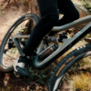 Nahaufnahme eines Mountainbikes: Rahmen mit Hinterradfederung, Pedal und Schuh, Reifen auf staubigem, grasigem Weg mit Steinen