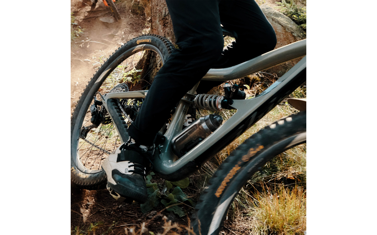 Nahaufnahme eines Mountainbikes: Rahmen mit Hinterradfederung, Pedal und Schuh, Reifen auf staubigem, grasigem Weg mit Steinen