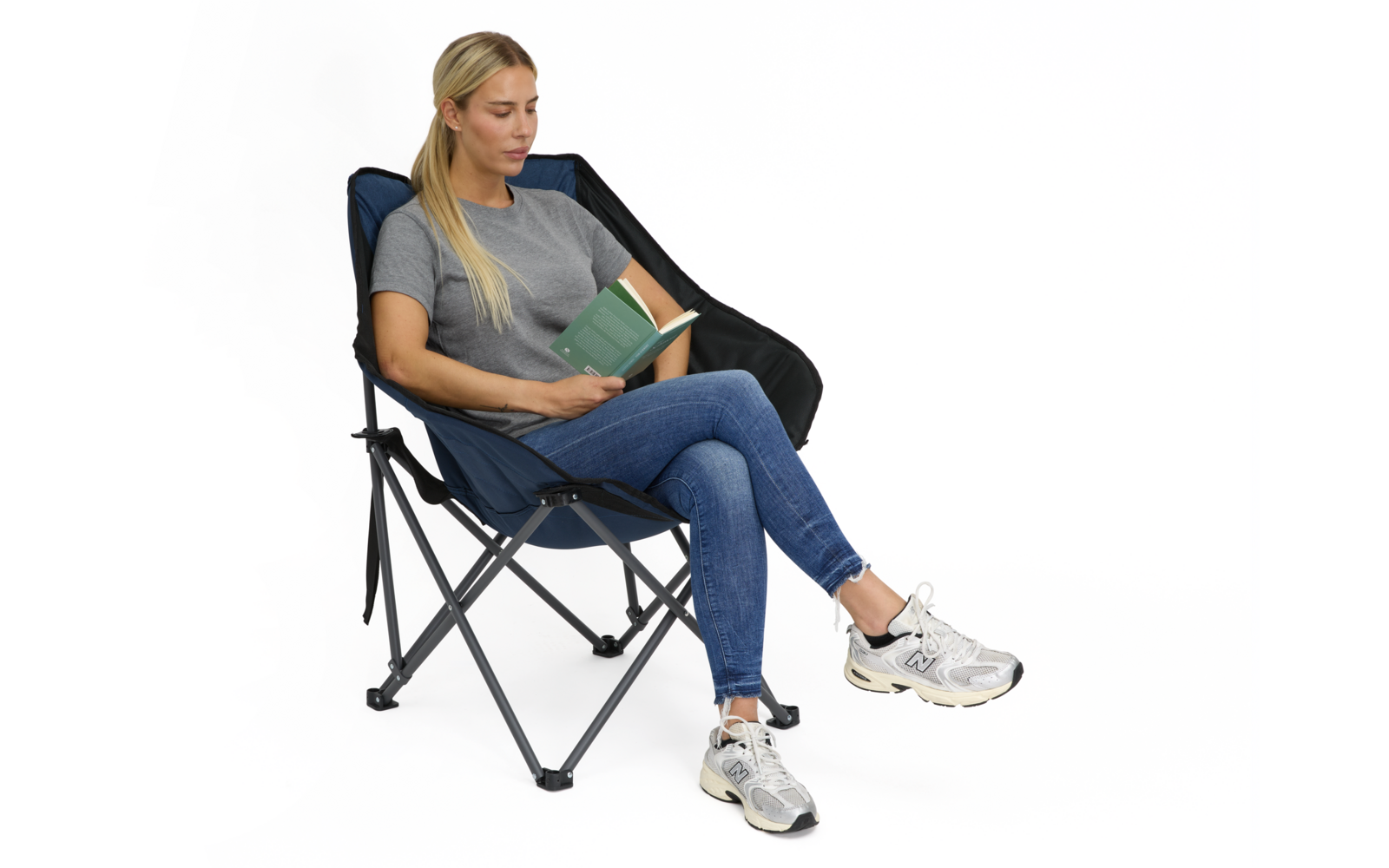Person sitzt in einem dunklen zusammenklappbaren Klappstuhl, Beine übereinander, hält ein geöffnetes grünes Buch, trägt graues T‑Shirt, blaue Jeans und weiße Sportschuhe, weißer Hintergrund