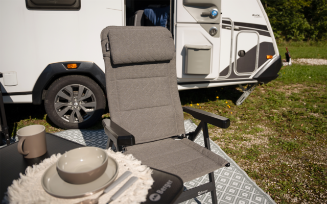Grauer Camping-Faltstuhl mit Armlehnen vor der Seitenwand eines weißen Wohnwagens; kleiner schwarzer Tisch mit beigeem Teller, Schüssel und Becher auf heller Stoffunterlage, gemusterte Bodenmatte sowie Gras und Bäume im Hintergrund