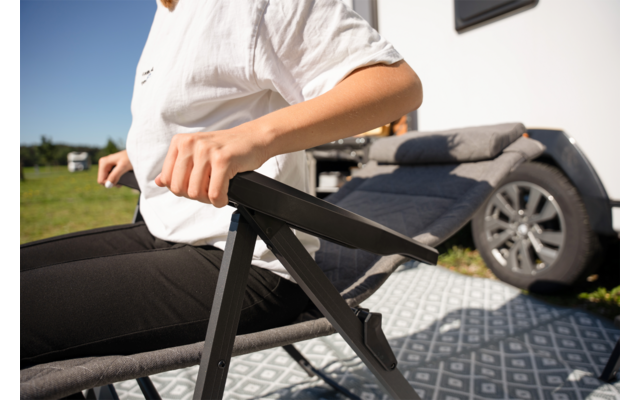 Oberkörper und Arme einer Person in weißem Shirt und schwarzer Hose auf einem schwarzen Klappstuhl neben einem Wohnwagen; Wohnwagenrad, gemusterte Bodenmatte, Rasen und blauer Himmel sichtbar