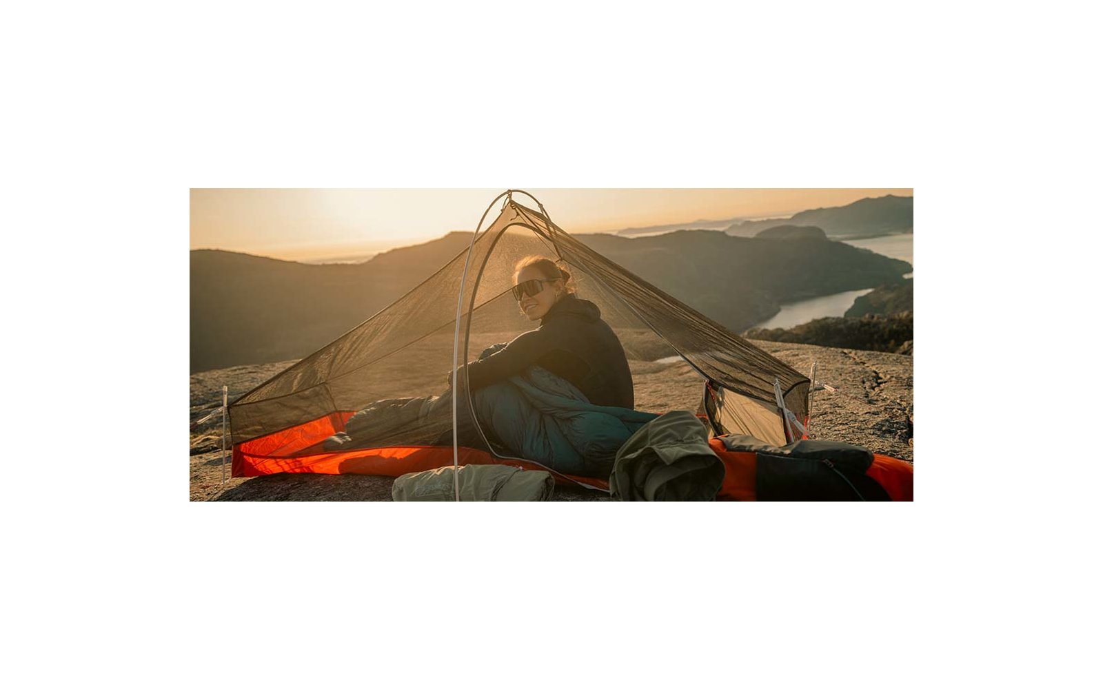 Persoon zit in een koepeltent met muskietennet op een slaapmat op een rotsrichel; meer en bergen op de achtergrond bij zonsondergang