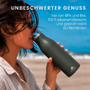 Frau im Profil trinkt aus dunkelgrüner Flasche am Strand; blauer Himmel und Meer im Hintergrund