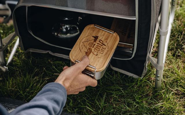 Hand sliding a metal box with bamboo lid and leather strap into a black fabric compartment under a folding chair; grass in the foreground
