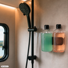 Zwarte handdouche met slang aan een rail, twee aan de muur gemonteerde doorzichtige dispenserflessen met groene en roze vloeistof, klein rechthoekig raam links