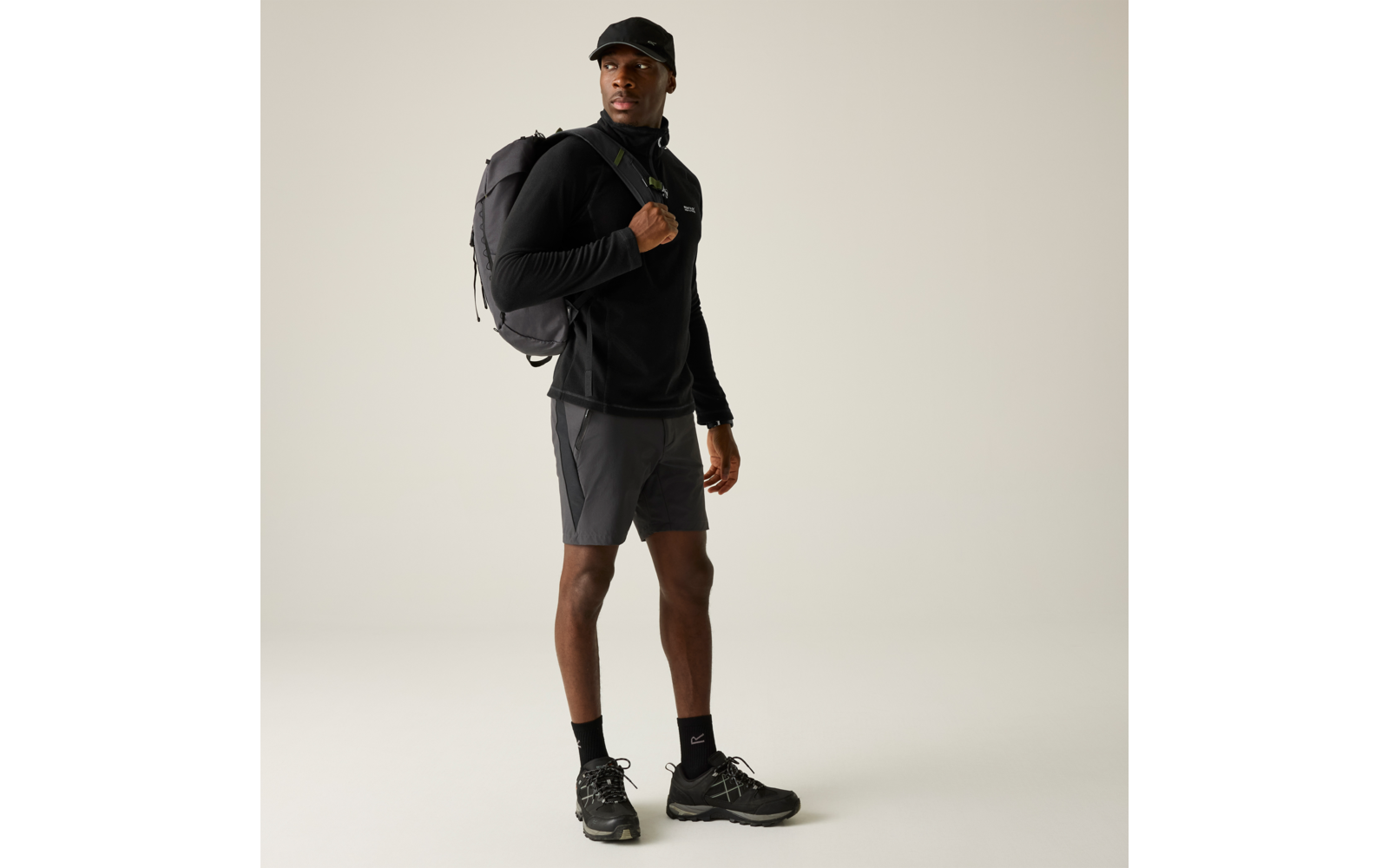 Person steht, trägt schwarze Kappe, schwarzen Pullover, graue Shorts, schwarze Socken und Sportschuhe, schwarzen Rucksack über einer Schulter