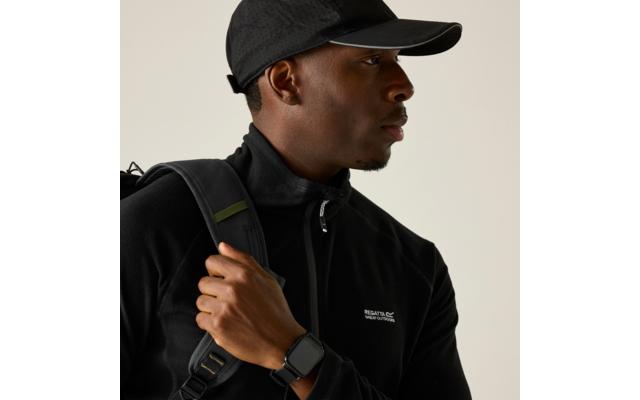 Person in seitlicher Ansicht, trägt schwarze Schirmmütze und schwarzes Oberteil mit Reißverschluss und kleinem weißen Logo, hält einen Rucksackriemen, trägt eine eckige Smartwatch am Handgelenk, heller einfarbiger Hintergrund