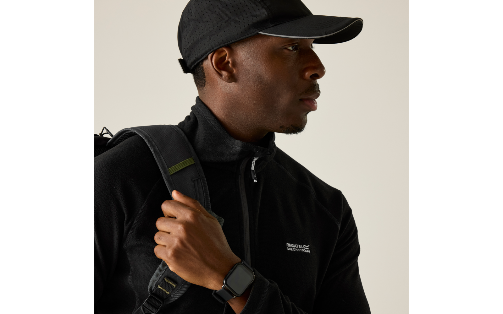 Person in seitlicher Ansicht, trägt schwarze Schirmmütze und schwarzes Oberteil mit Reißverschluss und kleinem weißen Logo, hält einen Rucksackriemen, trägt eine eckige Smartwatch am Handgelenk, heller einfarbiger Hintergrund