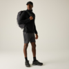 Person steht, trägt schwarze Kappe, schwarzen Pullover, graue Shorts, schwarze Socken und Sportschuhe, schwarzen Rucksack über einer Schulter