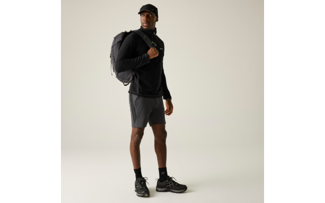 Person steht, trägt schwarze Kappe, schwarzen Pullover, graue Shorts, schwarze Socken und Sportschuhe, schwarzen Rucksack über einer Schulter