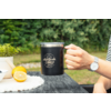 Main tenant une tasse noire avec le logo 'camp life' sur une table en bois; moitiés de citron, petit pot de plante et montre-bracelet visibles; arrière-plan végétal flou