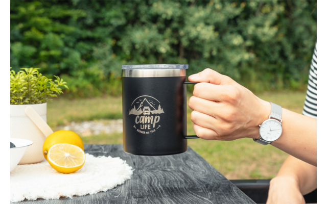 Main tenant une tasse noire avec le logo 'camp life' sur une table en bois; moitiés de citron, petit pot de plante et montre-bracelet visibles; arrière-plan végétal flou