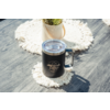 Tasse isolée noire avec couvercle transparent et logo "Camp Life", anse à droite, sur un dessous de tasse rond blanc en crochet posé sur une surface en bois grise; en arrière-plan un pot avec une succulente sur un autre dessous