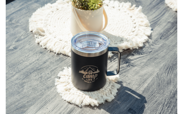 Tasse isolée noire avec couvercle transparent et logo "Camp Life", anse à droite, sur un dessous de tasse rond blanc en crochet posé sur une surface en bois grise; en arrière-plan un pot avec une succulente sur un autre dessous