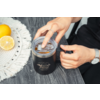 Deux mains ouvrant une tasse noire avec couvercle transparent et logo sur une surface en bois grise; à gauche un napperon rond blanc avec un citron entier et un citron coupé en deux; une main porte une bague, l'autre une montre-bracelet