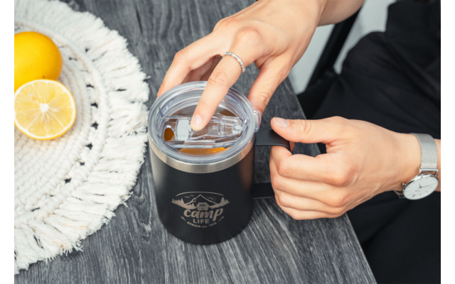 Deux mains ouvrant une tasse noire avec couvercle transparent et logo sur une surface en bois grise; à gauche un napperon rond blanc avec un citron entier et un citron coupé en deux; une main porte une bague, l'autre une montre-bracelet