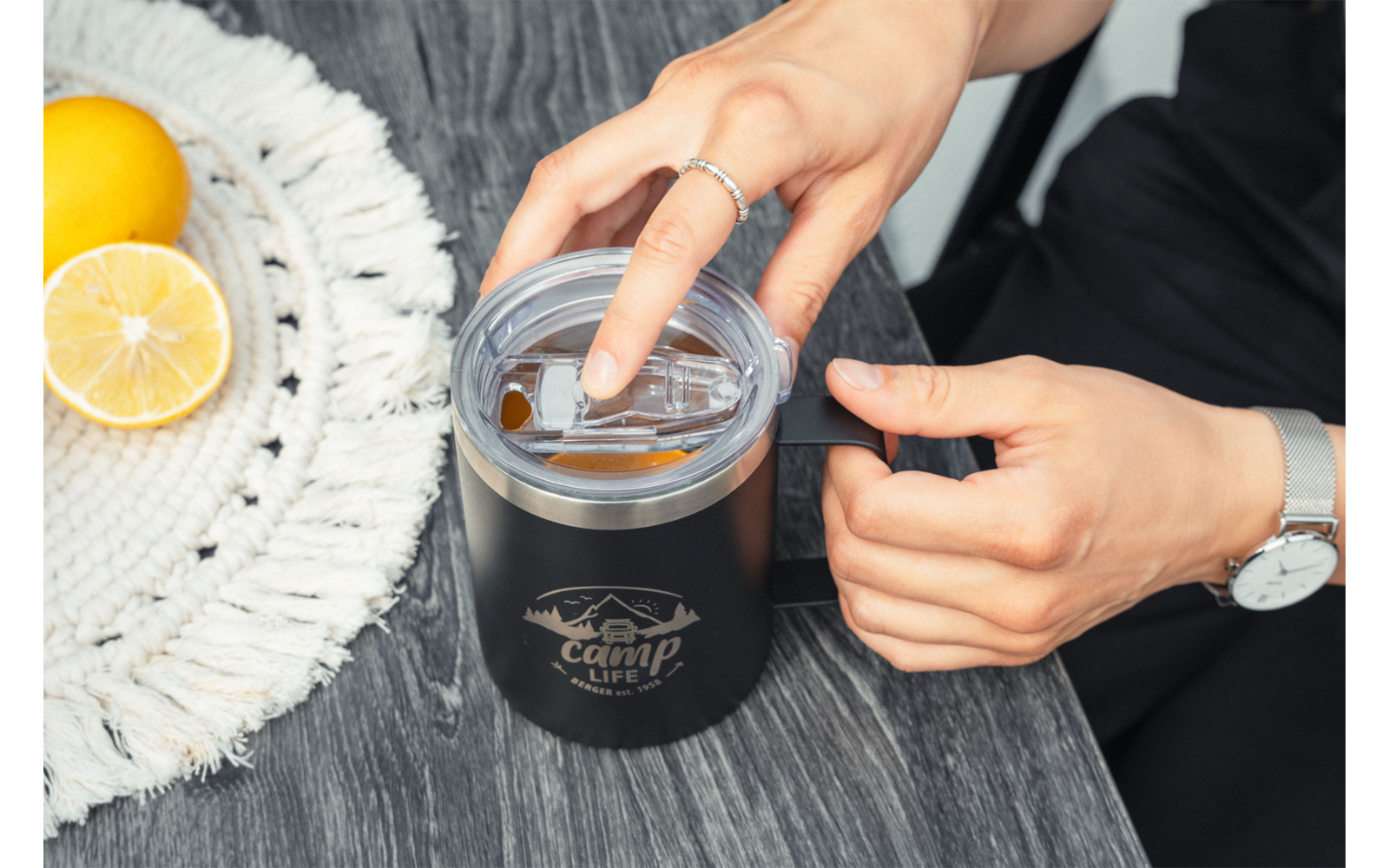 Deux mains ouvrant une tasse noire avec couvercle transparent et logo sur une surface en bois grise; à gauche un napperon rond blanc avec un citron entier et un citron coupé en deux; une main porte une bague, l'autre une montre-bracelet