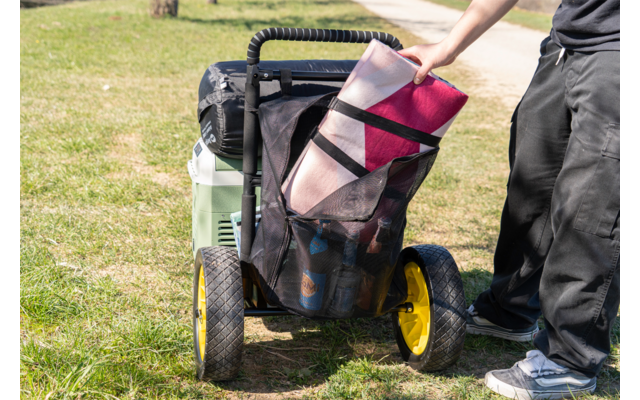 Mano coloca una manta doblada en una bolsa de malla en un carro de dos ruedas con ruedas amarillas; nevera verde en el carro; botellas visibles en la bolsa; persona con pantalones oscuros y zapatillas; césped y un camino al fondo